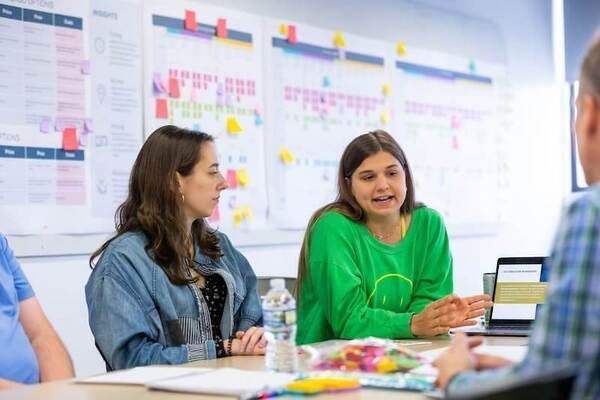 Two female students sitting with a group of people in a classroom for a design principles workshop.