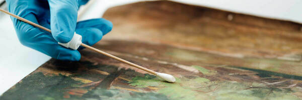 A gloved hand delicately cleans a painting with a cotton swab. The painting's colors are muted greens and browns, suggesting a natural landscape. This highlights the meticulous process of art restoration.