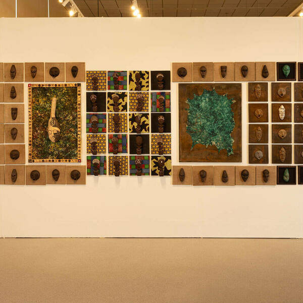 Wall display featuring a variety of artwork. Small sculpted heads are mounted on individual square canvases, arranged in grids.  A central artwork depicts a long, textured sculpture on a green and gold patterned background.  Another piece features a textured, teal abstract form on a rusted metal background.  Several small, framed squares contain additional sculpted heads and objects.