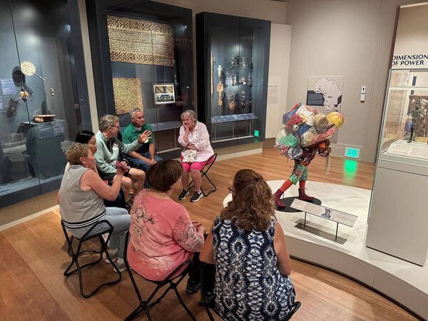 Adults seated in a museum gallery listen to a gesturing man. Nearby, a vibrant sculpture depicts a figure carrying a netted load.