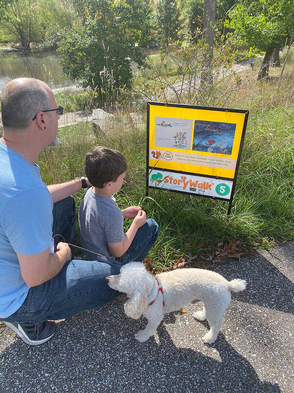 Man, boy, and white dog look at a yellow StoryWalk sign number 5 by a pond on a sunny day.